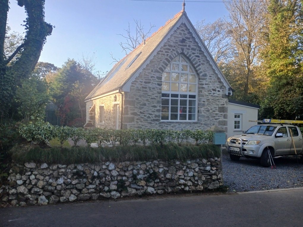 Restored stone building with a tall arched gable window, in sunshine behind a stone hedge
